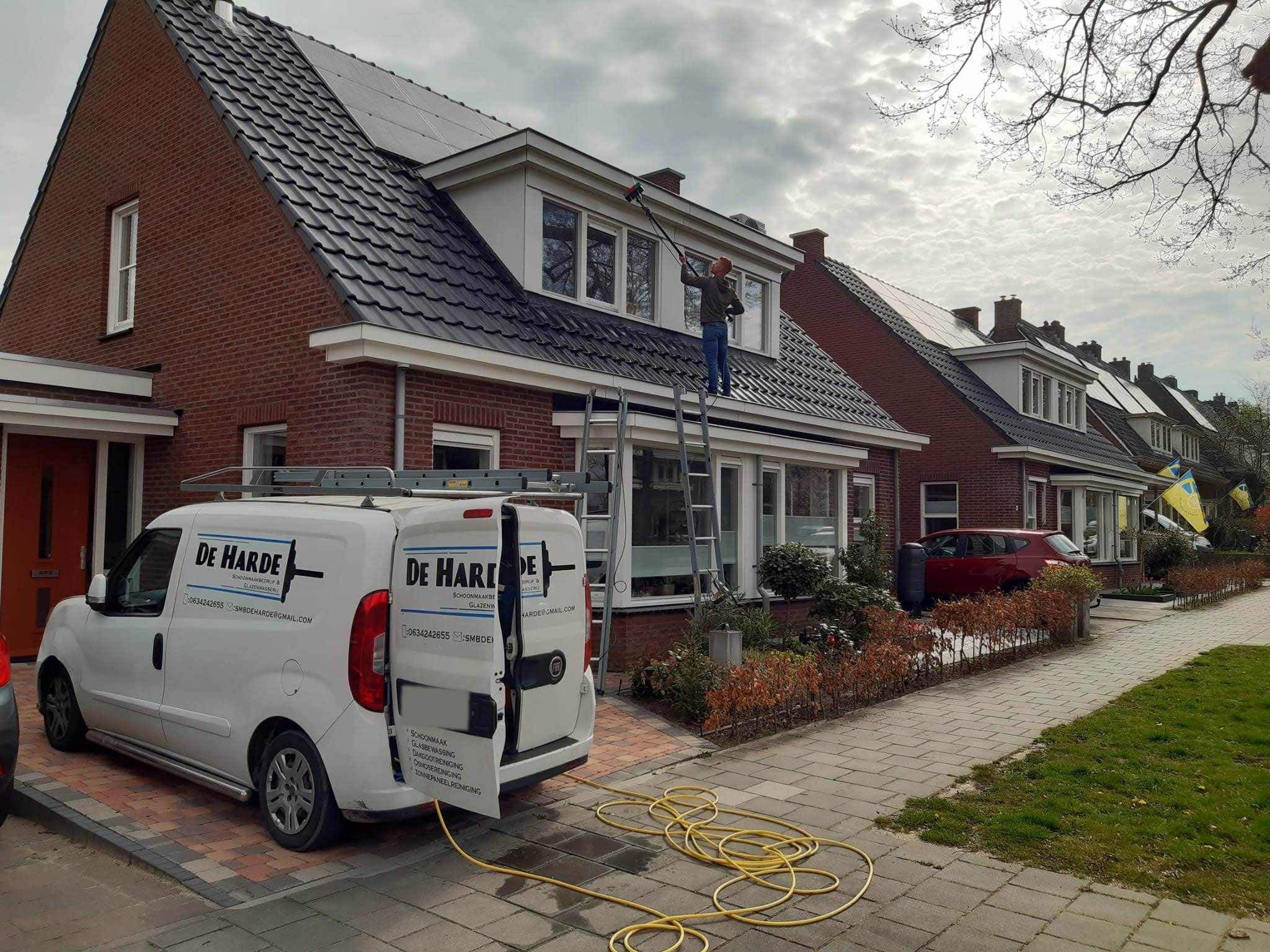 De bus van De Harde met open achterdeur waarop de diensten staan, voor een huis waar aan de dakkapel gewerkt wordt
